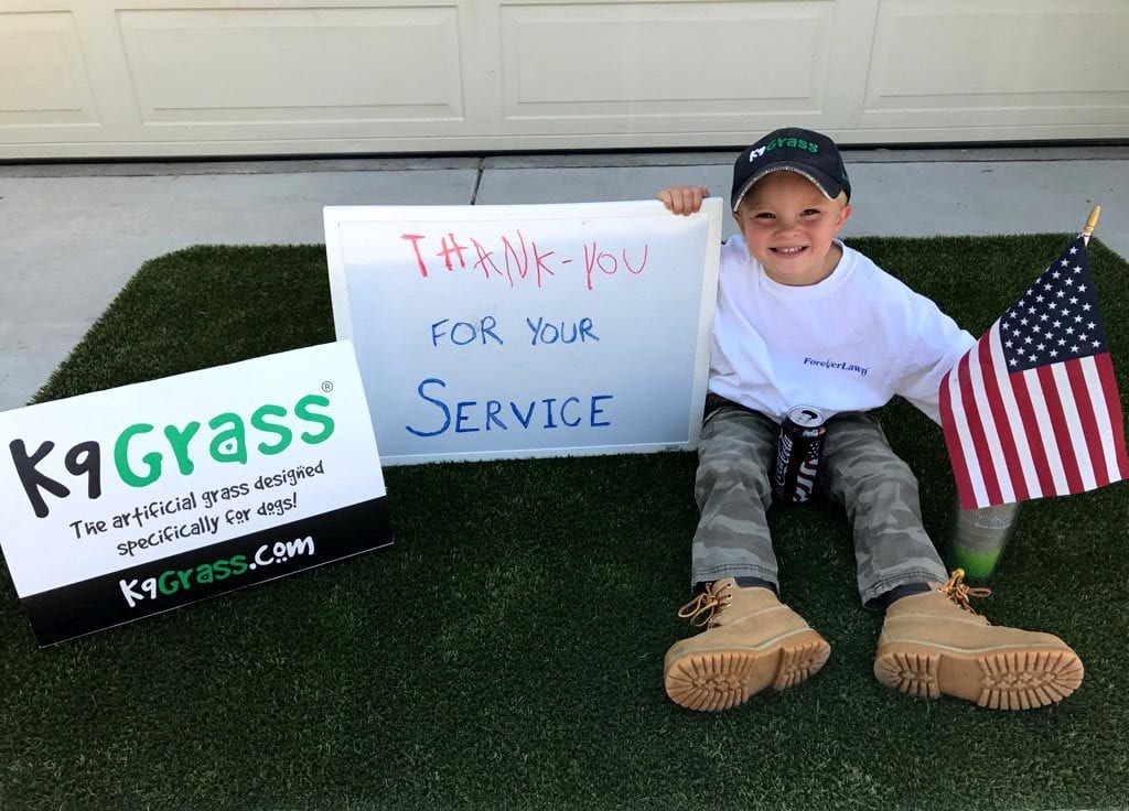 Boy sitting on K9Grass with sign for veterans.